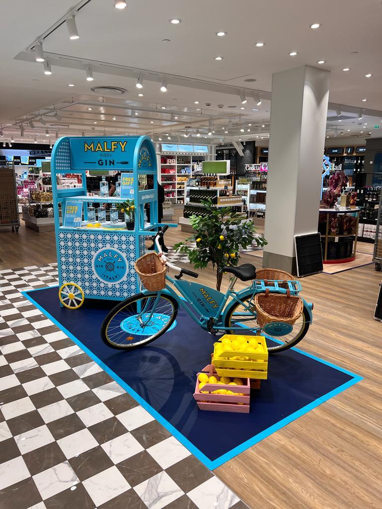 Vue d'ensemble du pop-up Malfy Gin : chariot, vélo et cagettes de citrons sur tapis bleu