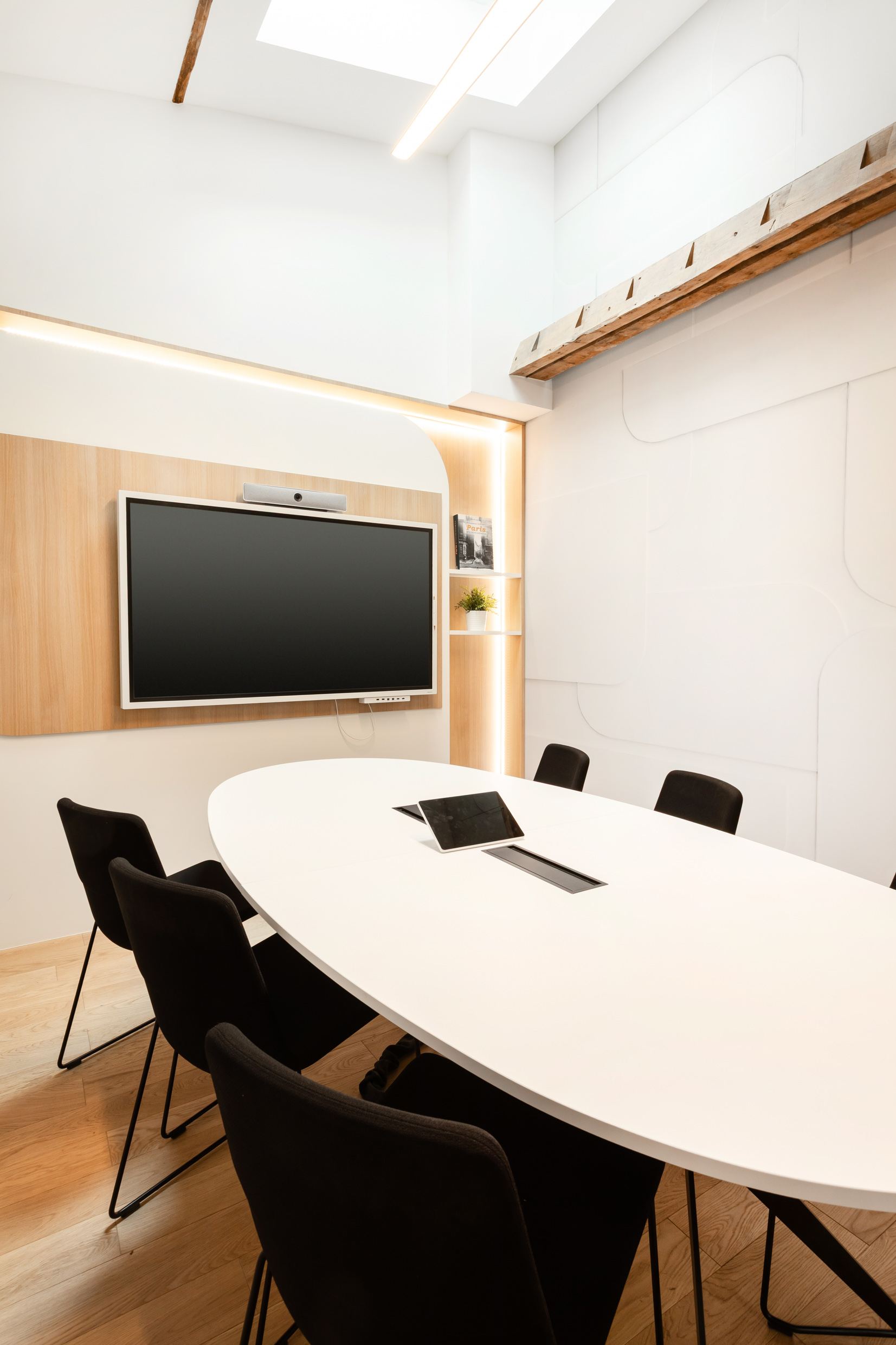 Salle de réunion visio du 25 BM aménagée par Isospace : écran TV intégré sur panneau bois, table ovale blanche et chaises noires design