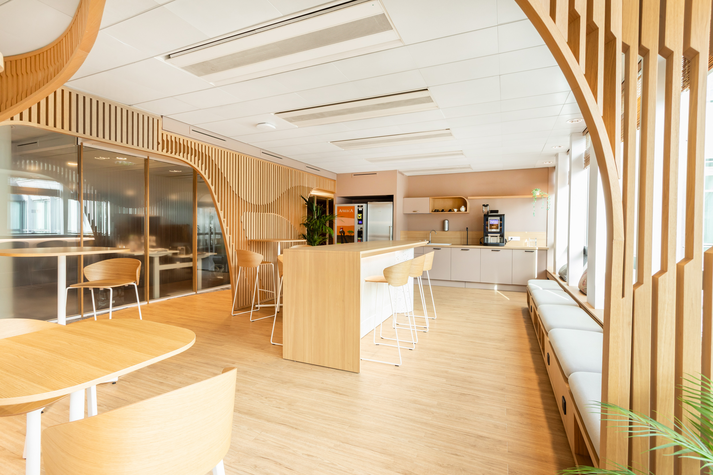Cuisine d'entreprise du 25 BM aménagée par Isospace : îlot central en bois clair, tabourets hauts, claustra bois et banquette blanche dans un siège social parisien