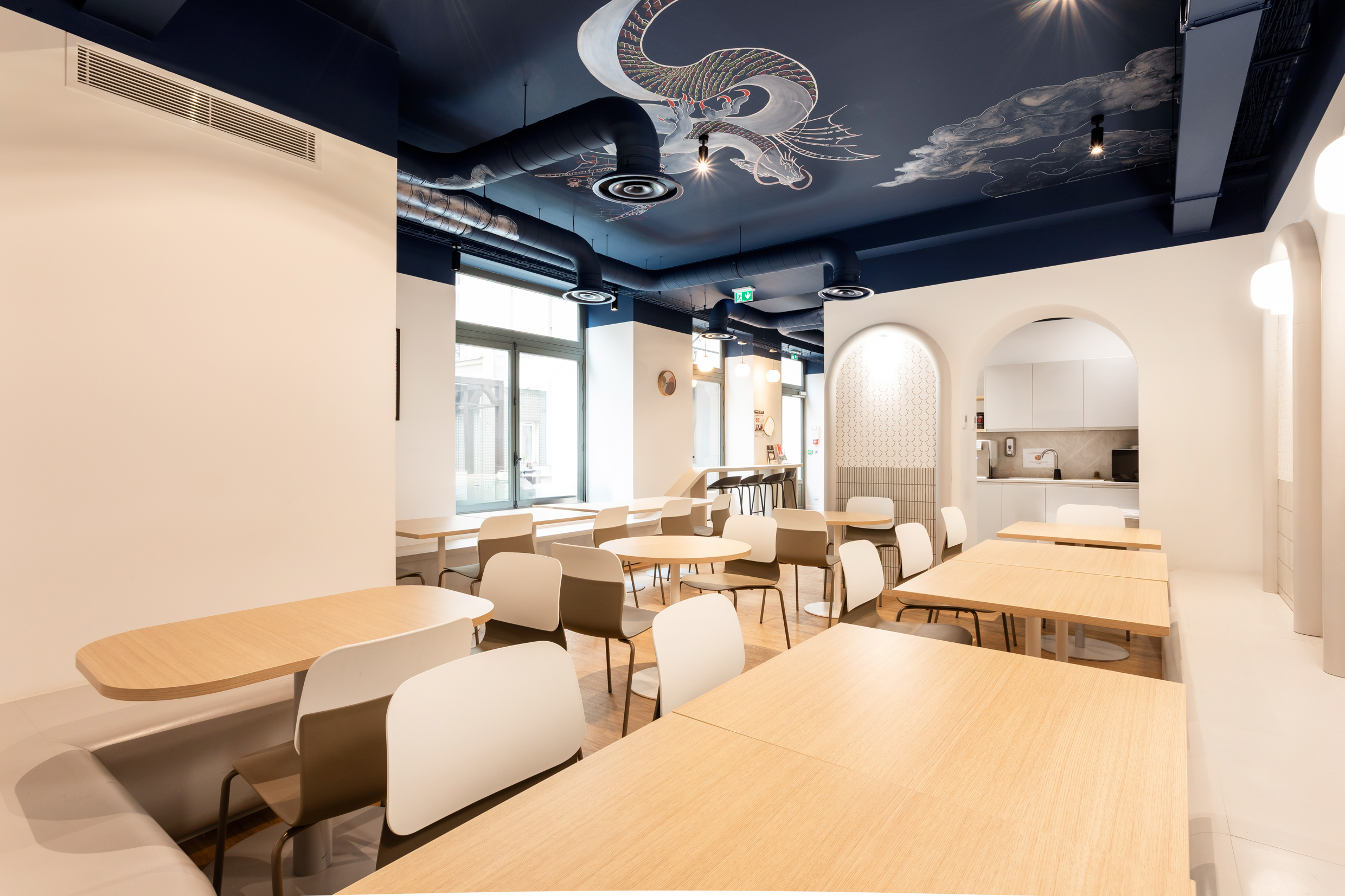 Cafétéria d'entreprise aménagée par Isospace : plafond bleu nuit avec fresque dragon, mobilier épuré bois clair, arche menant à la kitchenette