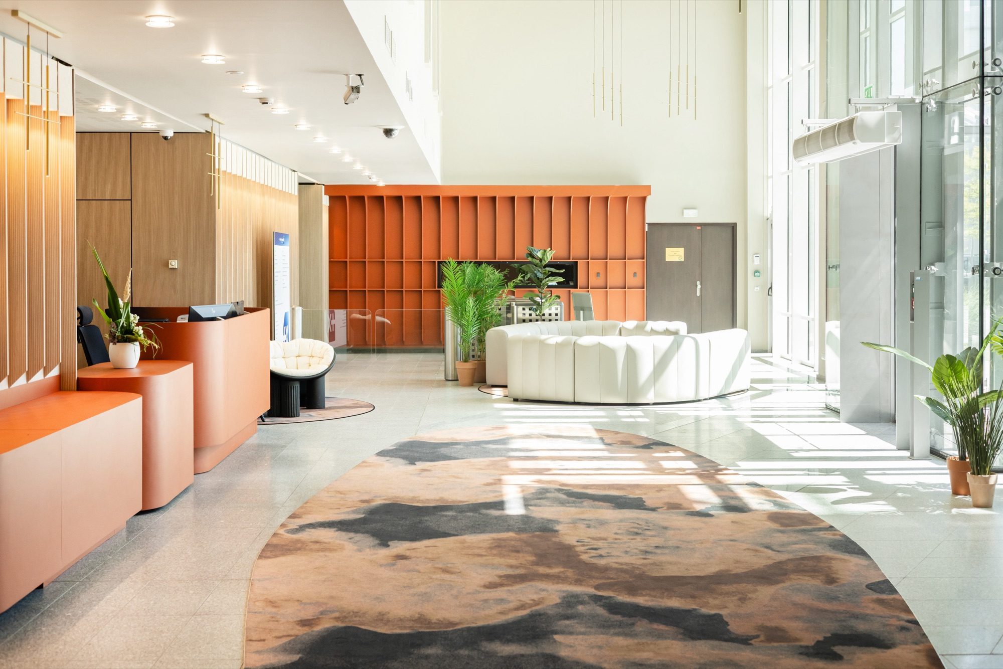 Hall d'accueil d'un siège tertiaire : mur d'arches orange, banque d'accueil sur mesure et tapis aux motifs organiques sur sol terrazzo