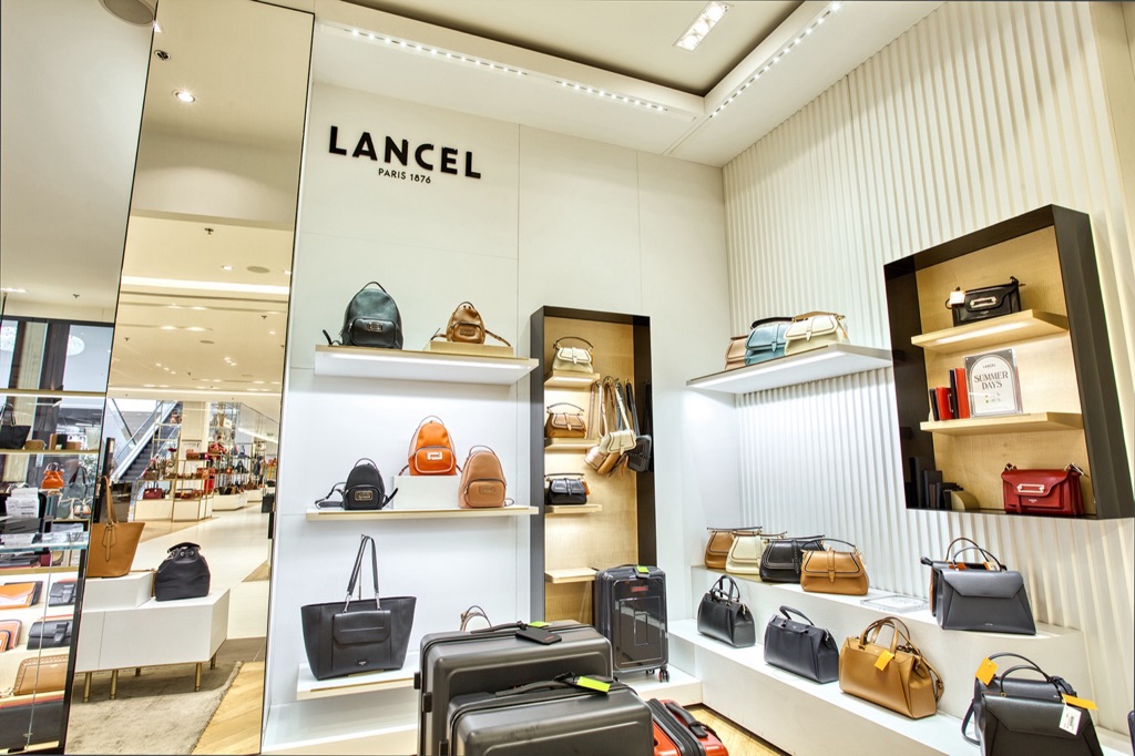 Meubles muraux blancs à étagères éclairées et niches noires présentant les sacs Lancel